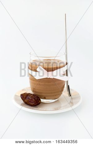 Homemade smoothie made out of bananas almond milk espresso and medjool dates in a glass with pink bow on white background. A drinking spoon and a medjool date on a cream colored saucer.