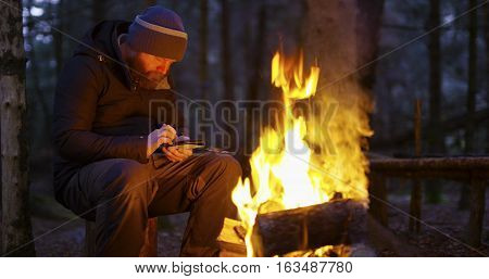 One focused man with beard warms himself by the campfire outdoors in the woods a cold winter evening. Uses a smart phone and compass in the forest.