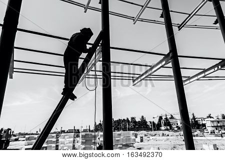 Silhouetted industrial shot of construction worker who is using the wooden ladder to welding metal frame.