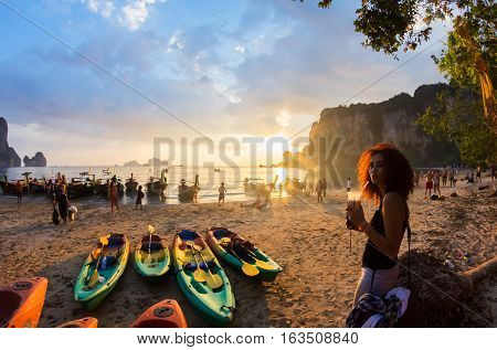 KRABI, THAILAND, 07 JANUARY 2012 : Young woman with a milkshake, kayaks and traditional thai longtail boats at the sunset, Tonsai beach near Railay bay