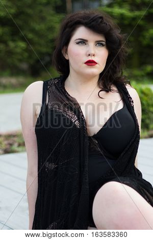 Young beautiful busty curvy plus size model with big breast in black bra and lace cape xxl woman professional makeup and hairstyle sitting outdoors