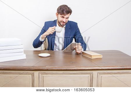 Technology people and business concept - handsome man with beard and brown hair and blue suit and tablet pc computer and some books and cup drinking coffee or tea sitting in the office holding tablet and smiling. Isolated on white background.