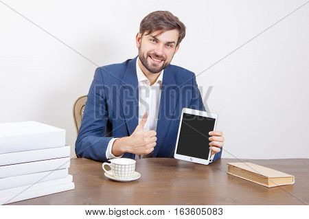 Technology people and business concept - handsome man with beard and brown hair and blue suit and tablet pc computer and some books in the office holding tablet and showing display screen with thumb. Isolated on white background.