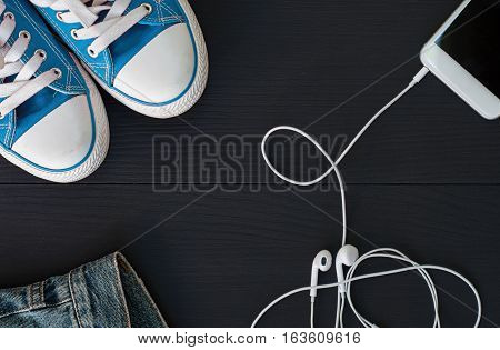 Smartphone with headphones and blue sneakers on a black wooden surface top view