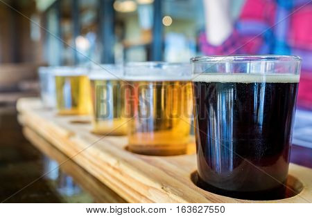 Beer samplers in small glasses individually placed in holes fashioned into a unique wooden tray. Focus is on the backlit front dark stout.