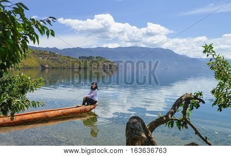 Single young Chinese woman smiles to the camera, sitting on boat by the bank of Lugu lake, Lijiang, China
