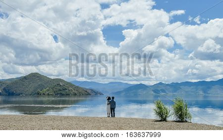 Young Chinese couple hold each other, standing hand in hand on bank of Lugu lake, back to camera,  at Daluoshui village, Lijiang, China