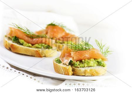 smoked salmon and avocado cream or guacamole on baguette slices on a white plate as a healthy party snack bright background copy space closeup