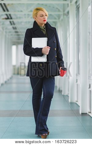 Portrait of blond businesswoman holding coffee and laptop in lobby.