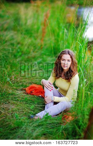 Young pensive girl sitting on the shore of the lake on the trampled green grass and enjoy the beauty of nature