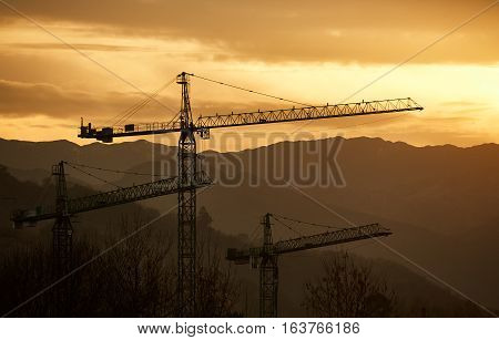 Three construction cranes in front of mountains during sunset