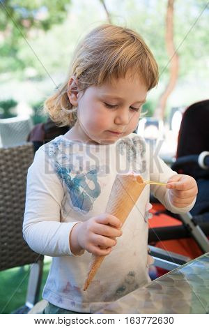 portrait of two years old blonde cute child with white shirt eating strawberry ice cream cone with spoon at urban park