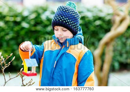 Little kid boy feeding birds in winter. Cute happy preschool child hanging colorful selfmade bird house on tree on frosty cold day. Preschooler in colorful wam clothes. Nature, empathy with animals.