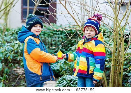 Two little kids boys feeding birds in winter. Cute happy preschool children, siblings and friends hanging colorful selfmade bird house on tree on frosty cold day. Preschoolers in colorful wam clothes.