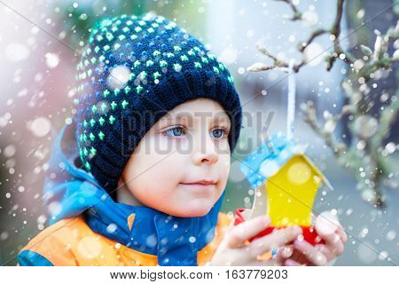 Little kid feeding birds in winter. Child hanging colorful selfmade bird house on tree on frosty cold day. Toddler in colorful wam clothes. Selective focus on child and feeder with seeds