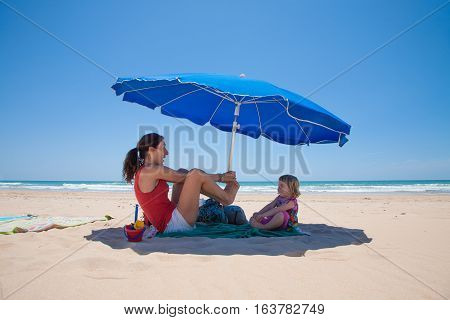 two years old blonde girl and mother red shirt playing and smiling happy on green towel over sand beach in shade down of blue parasol with ocean behind