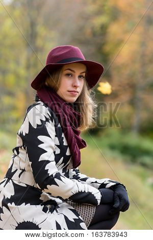 Woman in a floral patterned coat and wine red hat in the park. Happy girl and colorful autumn forest. Portrait of a lady yellow green red and brown leaves background