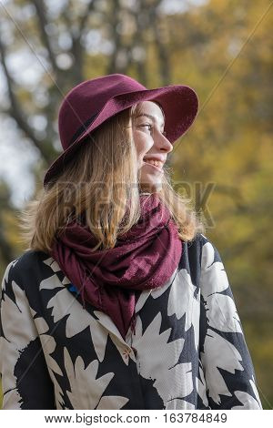 Woman in a floral patterned coat and wine red hat in the park. Happy girl and colorful autumn forest. Portrait of a lady yellow green red and brown leaves background
