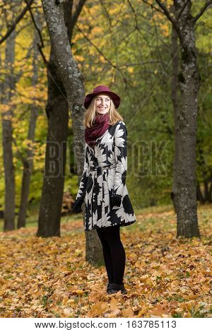 Woman in a floral patterned coat and wine red hat in the park. Happy girl and colorful autumn forest. Portrait of a lady yellow green red and brown leaves background