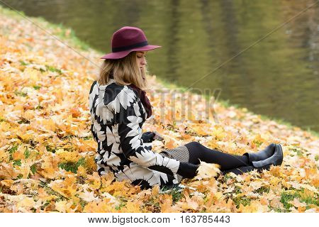 Woman in a floral patterned coat and wine red hat in the park by the river. Happy girl and colorful autumn forest. Portrait of a lady yellow green red and brown leaves background