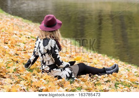 Woman in a floral patterned coat and wine red hat in the park by the river. Happy girl and colorful autumn forest. Portrait of a lady yellow green red and brown leaves background
