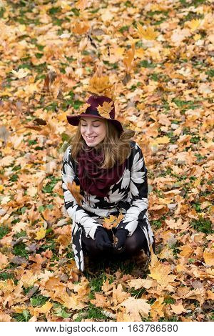 Woman in a floral patterned coat and wine red hat playing with leaves in the park. Happy girl throws maple tree leaves into the air. Yellow green red and brown leaves colorful autumn forest