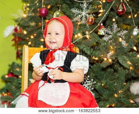 Portrait of girl child in suit a red hat for Christmas around a fir-tree decorated. Kid on holiday new year