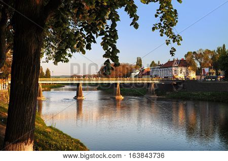 View on the main bridge across the river Uzh in Uzhhorod in warm sunset light. High green tree growing nearby its branches hanging over the river