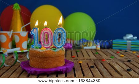 Birthday cake with candles on rustic wooden table with background of colorful balloons, gifts, plastic cups and candies with blue wall in the background. Focus is on cake