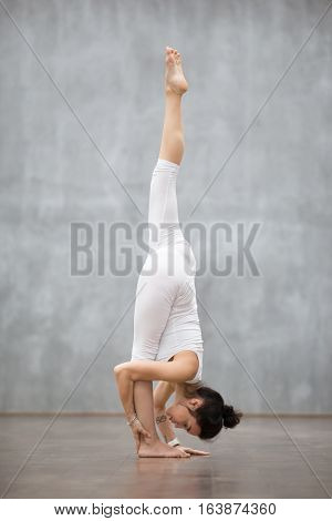 Profile portrait of beautiful young woman wearing white tank top working out against grey wall, doing yoga or pilates exercise. Standing splits, urdhva prasarita eka padasana. Full length