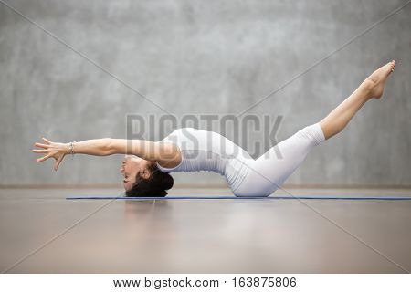 Side view portrait of beautiful young woman wearing white sportswear working out in fitness center against grey wall, doing yoga or pilates exercise. Variation of Matsyasana, Fish pose. Full length