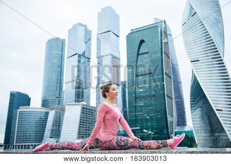 Young attractive woman practicing yoga, sitting in Monkey God exercise, Splits, Hanumanasana pose, working out wearing pink sportswear, outdoor full length, modern urban skyscraper background