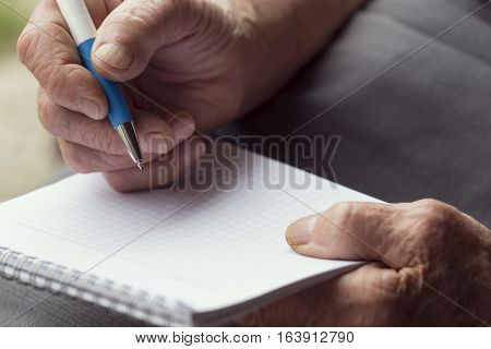 Detail of an elderly woman's hands holding a pen and writing a grocery list. Selective focus