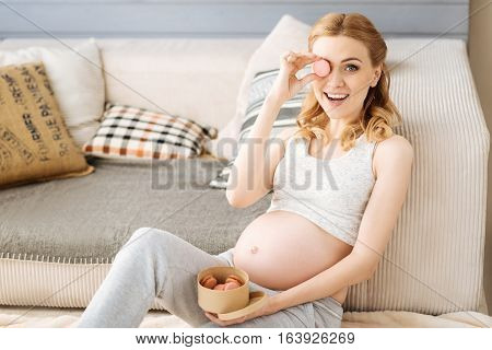 Look how funny. Pregnant young delighted woman posing with macaroons while sitting on a sofa and enjoying leisure time.