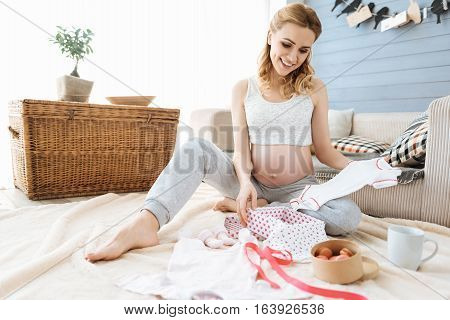 The cutest thing. Pregnant delighted young woman enjoying new baby clothes while smiling and sitting in a cozy room.