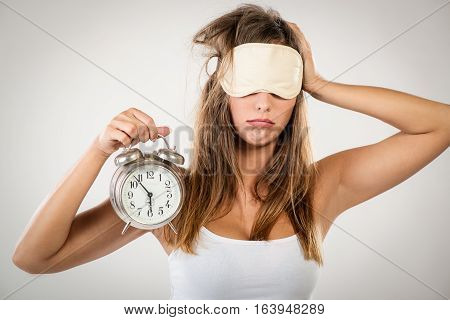 Beautiful young woman with sleeping mask holding alarm clock. She is tired and lazy in morning.