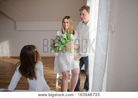 Young man congratulates his pregnant wife. She sits in a beautiful white dress with a bouquet of tulips. Husband gently hugs a wife and lovingly looks at her. Little daughter looks at the parents.