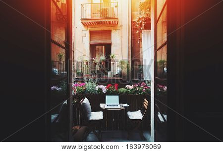 Small cozy beautiful workspace on balcony with laptop mouse smartphone two chairs and flowers around on sunny summer day seeing through opened doors from dark interior facade of house behind