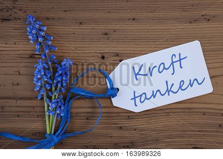 Label With German Text Kraft Tanken Means Relax. Blue Spring Grape Hyacinth With Ribbon. Aged, Rustic Wodden Background. Greeting Card For Spring Season