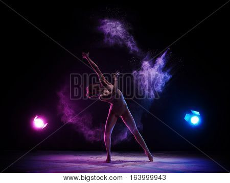 Female sexy gymnast in beige bodysuit bending gracefully with her hands up on gray background with colored dust all over body shot made with color filter.