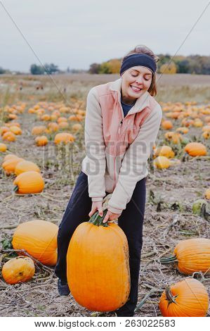 Adult Woman (30s) Attempts To Pick Up A Giant Pumpkin From A Pumpkin Patch. Smiling And Laughing And