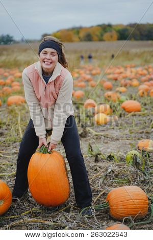 Adult Woman (30s) Attempts To Pick Up A Giant Pumpkin From A Pumpkin Patch. Smiling And Laughing And