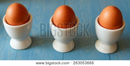 3 Boiled Eggs In White Egg Cups On A Blue Wooden Background