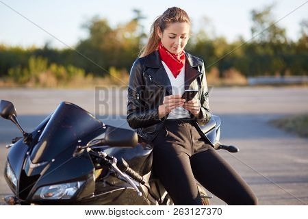 Photo Of Female Driver Holds Smart Phone In Hands, Surfes Bikers Blog In Internet, Wears Stylish Clo