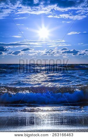Bright Blue Surf. Waves Crash Onto A Glistening Sandy Beach With A Low Angle Sunburst. Vertical Seas