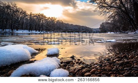 Winter River Sunset. As ice flows downstream, golden sunlight reflects off the water and snow along this peaceful riverside. Seasonal landscape background with Copy Space.