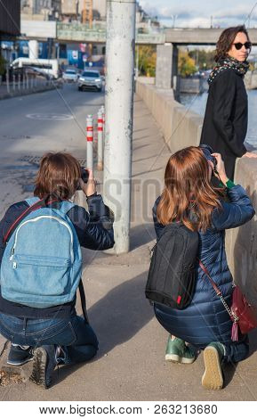 Photographer In The Street Photo Shooting Process. Model Is Young Lady In Stylish Black Coat With Bl
