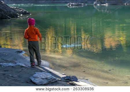 A Little Girl, A Child, In Warm Clothes And A Red Cap Throws Stones Into The Water From The Sandy Sh
