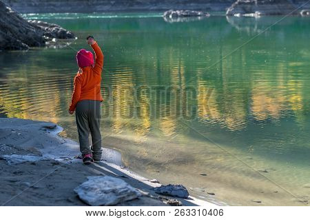 A Little Girl, A Child, In Warm Clothes And A Red Cap Throws Stones Into The Water From The Sandy Sh