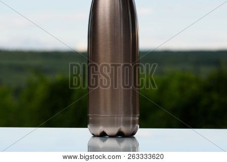 Thermo Stainless Bottle For Water, Tea Or Coffee. Sky And Forest On Background. On The Glass Desk. T